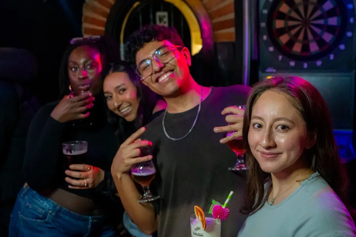 Grupo de cuatro personas sonriendo y sosteniendo bebidas en un bar con un tablero de dardos en el fondo.