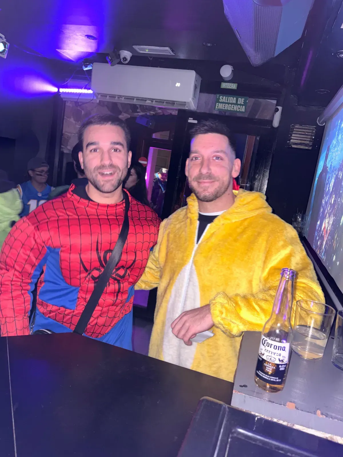 Dos hombres disfrazados en un bar: Spider-Man y un pájaro amarillo. Cerveza y bebidas en la barra.