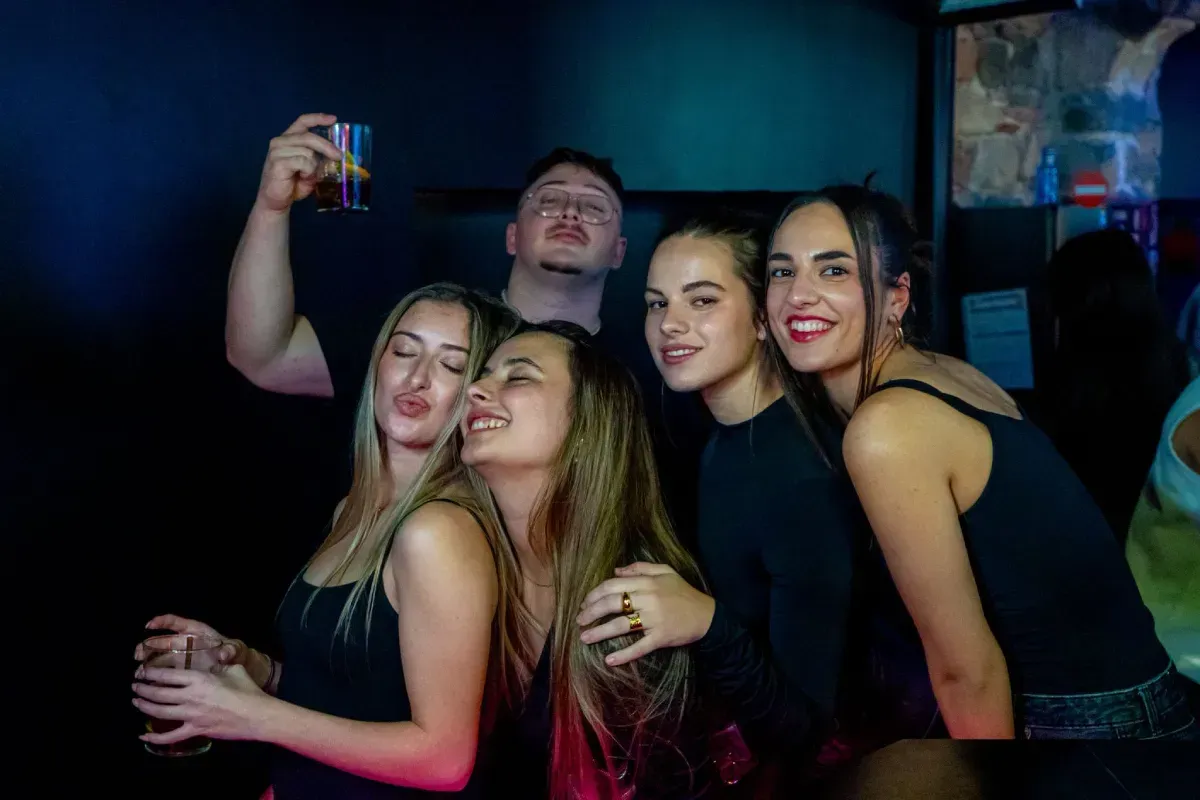 Grupo de amigos en un bar oscuro, sonriendo y riendo. Uno de ellos levanta una copa, celebrando.
