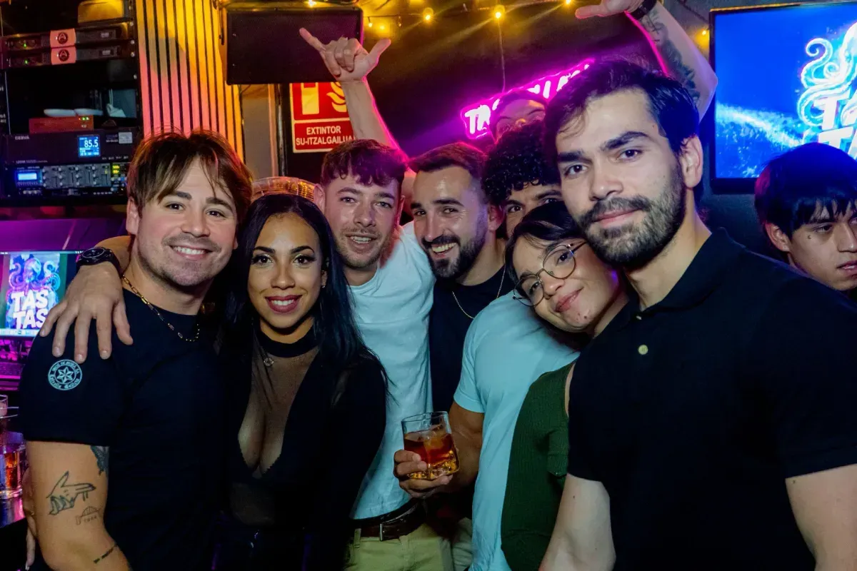 Grupo de personas sonriendo en un bar. Algunos se abrazan. Ambiente festivo con luces de colores.