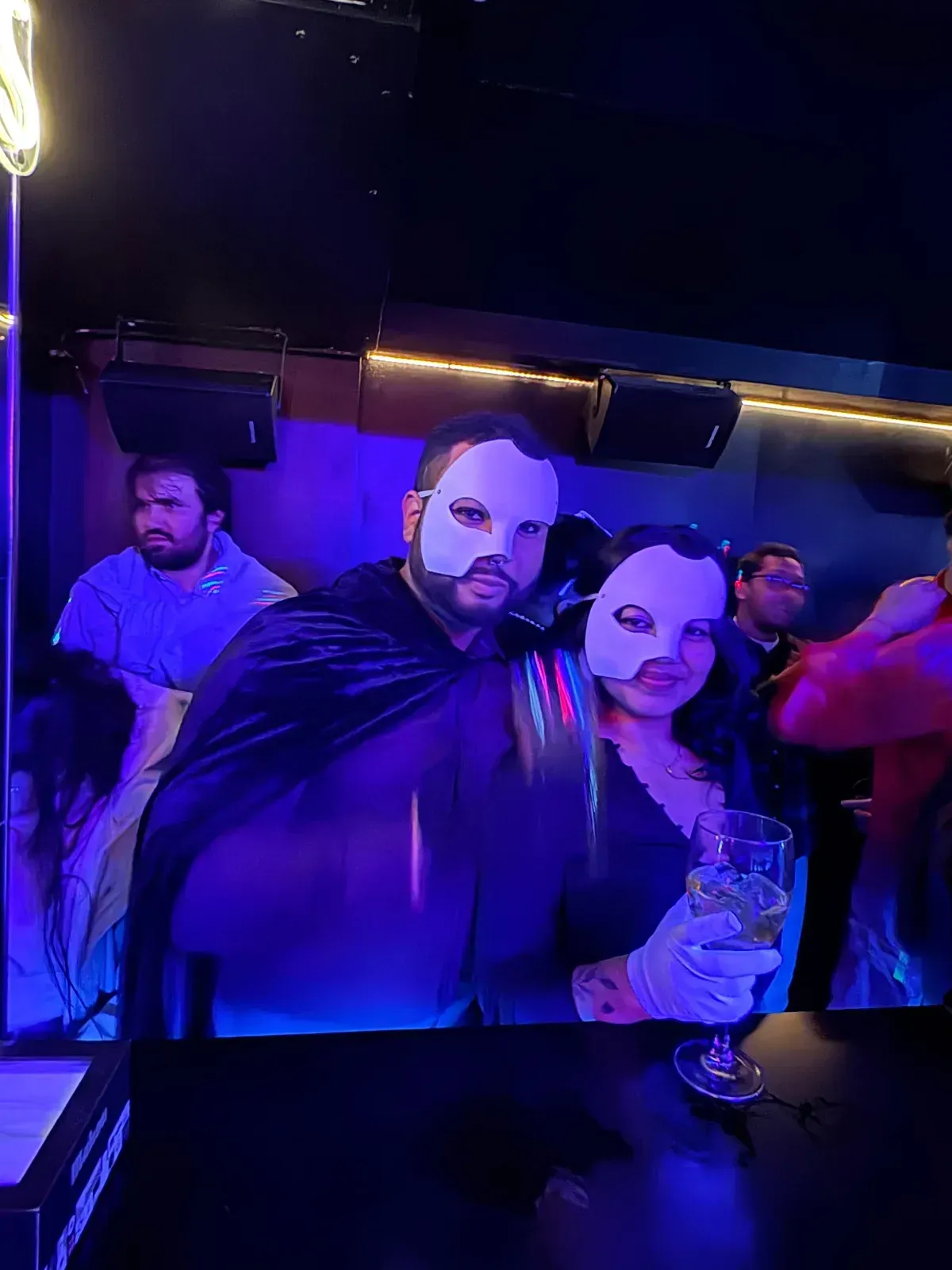 Una pareja con máscaras de El Fantasma de la Ópera posa en un bar. Uno lleva una capa negra. Se ven otros al fondo.