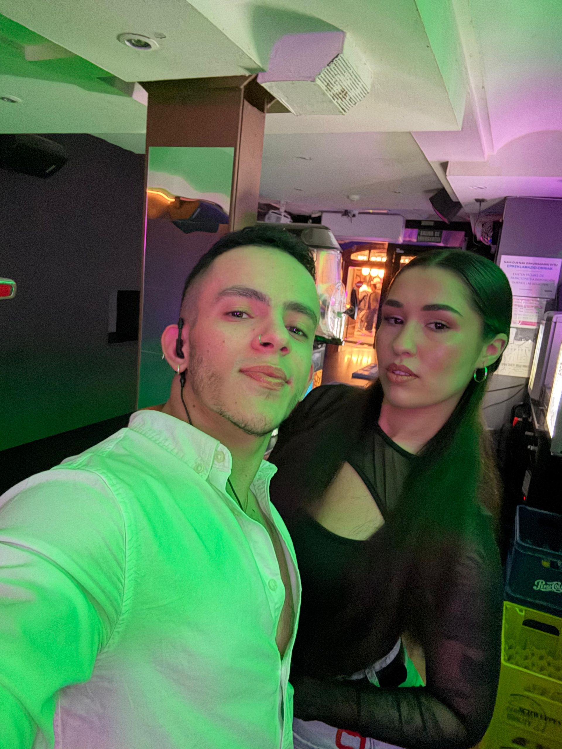 Dos personas posando en un local interior iluminado con luces de neón, una con una camisa blanca y la otra con una blusa negra.