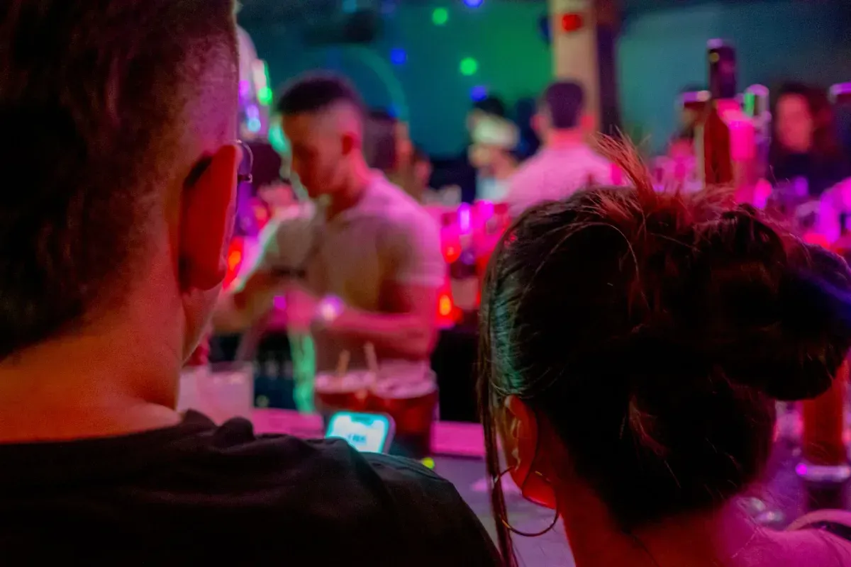 Gente en un bar mirando a un camarero. Luces brillantes y coloridas iluminan la escena.