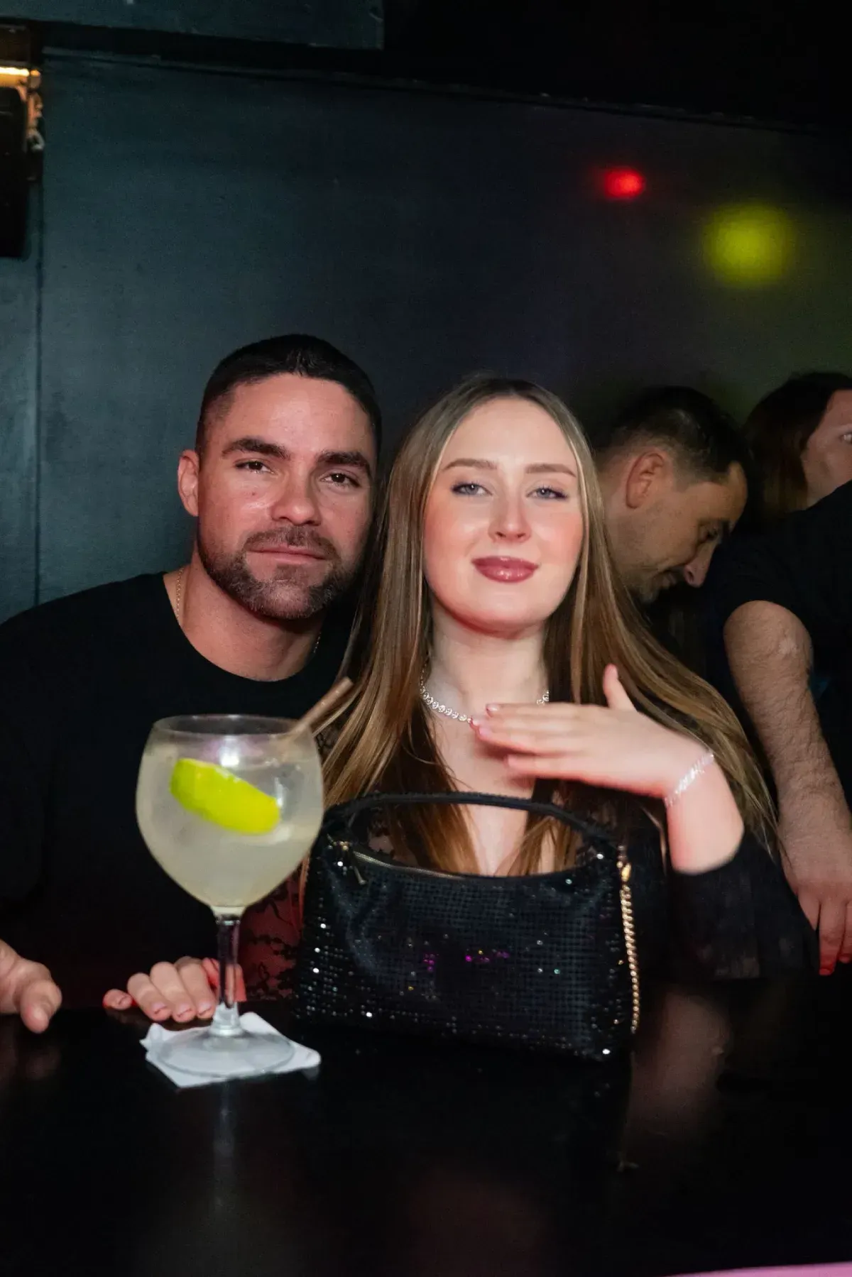 Hombre y mujer en un bar, mujer señalando, cóctel en el mostrador, entorno oscuro.