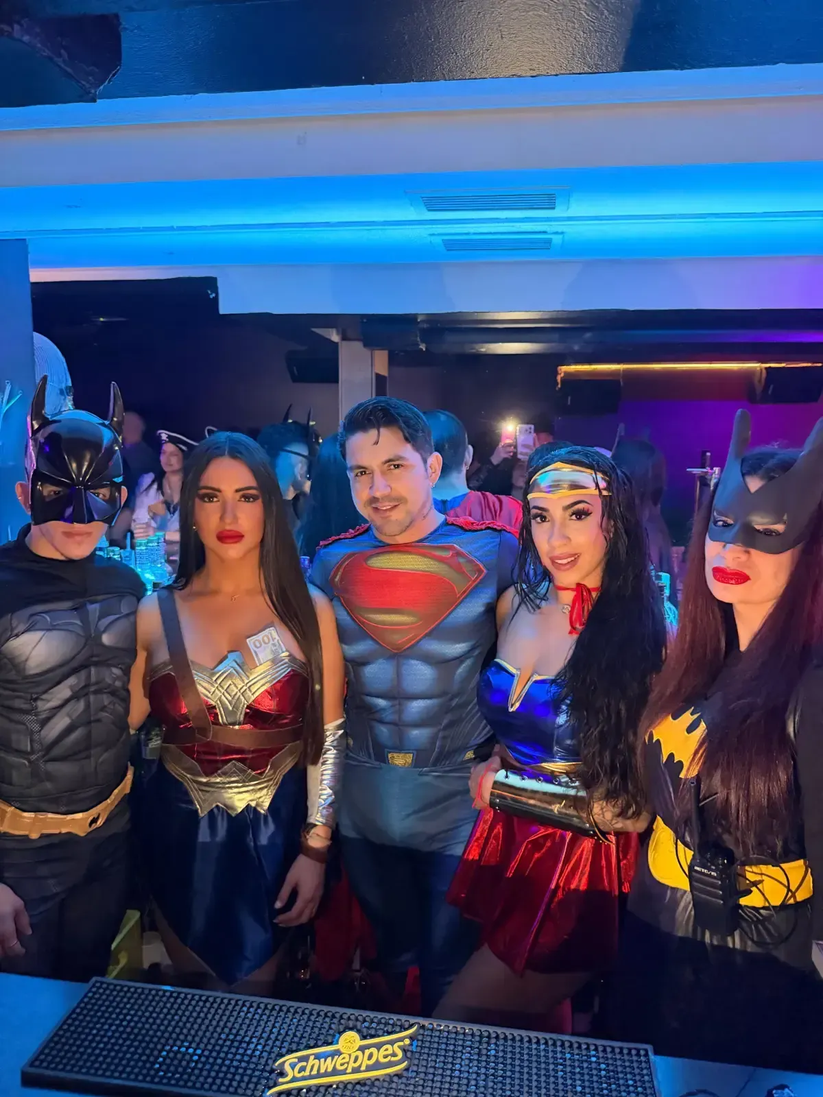 Cinco personas disfrazadas de superhéroes posan en un bar. Batman, la Mujer Maravilla, Superman y Batgirl están cerca.