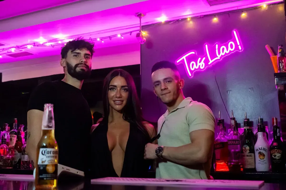 Tres personas en un bar frente a un letrero de neón. Dos hombres y una mujer posan con bebidas, iluminados por una luz rosa.