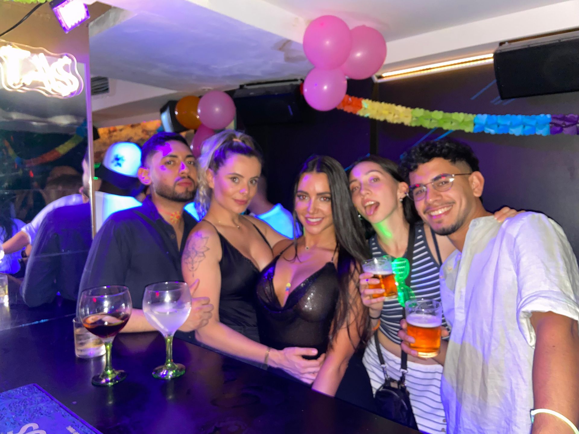 Cinco amigos sonriendo en un bar, con cócteles y globos rosas en un ambiente de fiesta con luz tenue.