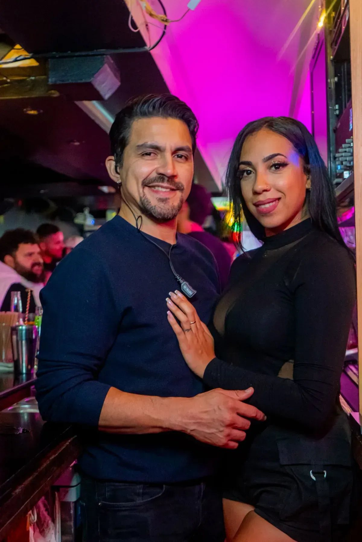 Un hombre y una mujer sonríen en una barra, apoyados en ella. Están rodeados de otras personas.