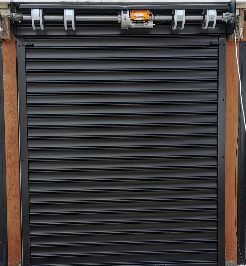 Volet roulant en métal noir sur une façade de magasin, fermé par un mécanisme motorisé situé au-dessus