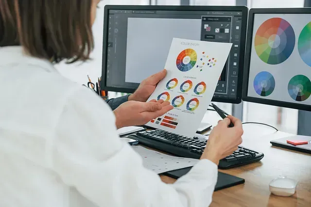 Una mujer está sentada en un escritorio frente a una computadora sosteniendo un trozo de papel.