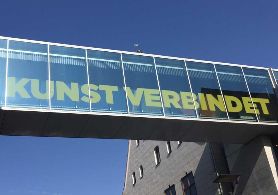 Eine gläserne Brücke über einem Gebäude mit der Aufschrift „Kunst verbindet“