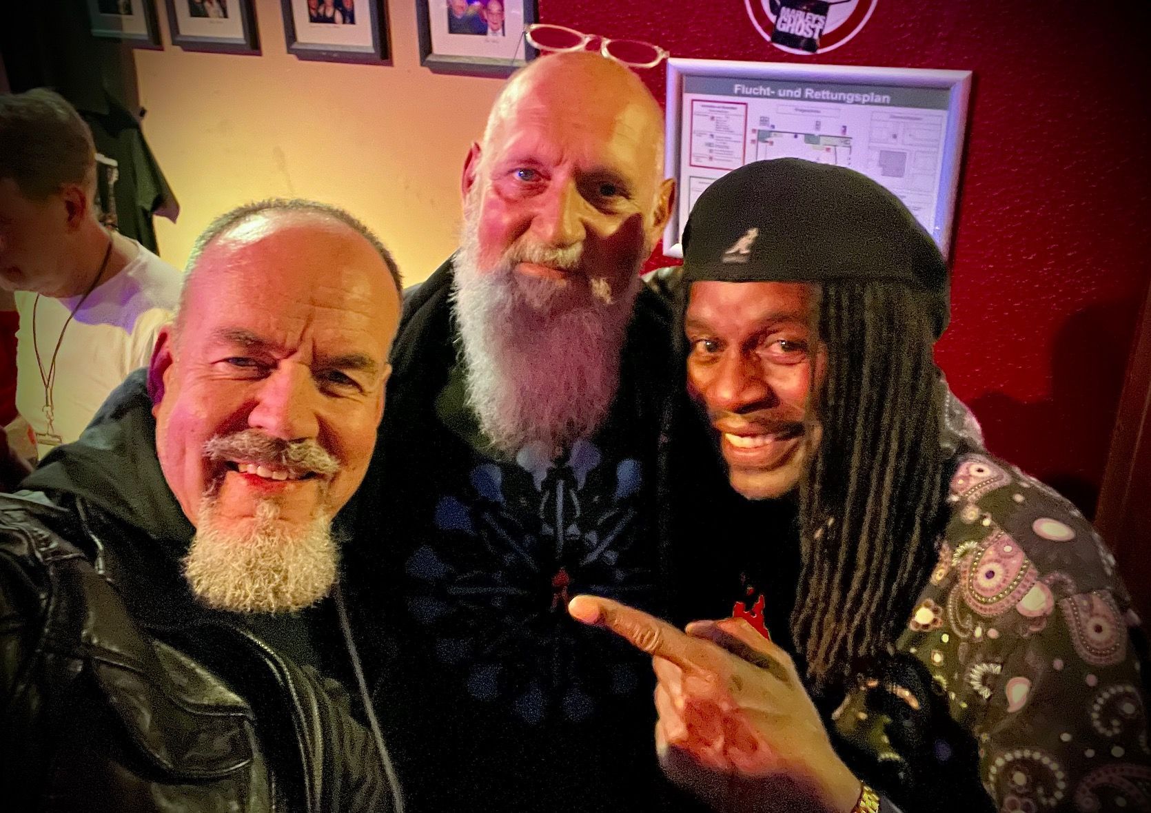 Drei Männer lächeln für ein Selfie in einem Innenraum; hinter ihnen hängen gerahmte Bilder an der Wand.