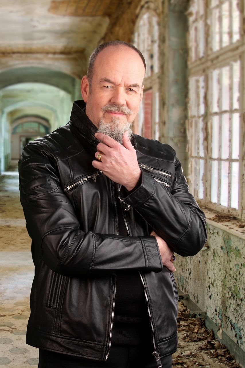 Eine Person mit Bart und dunkler Lederjacke steht in einem langen, heruntergekommenen Flur mit großen Fenstern.