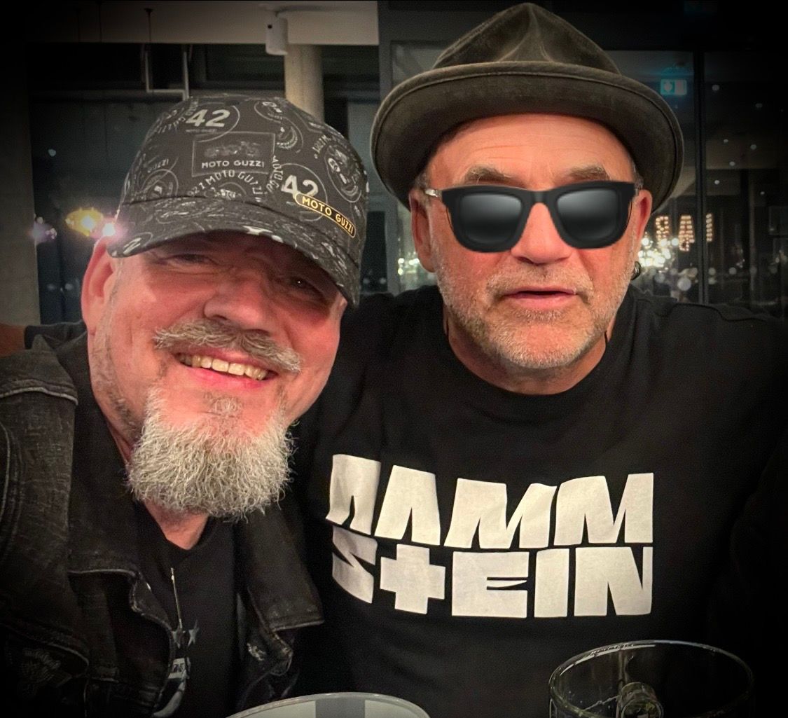 Zwei Männer lächeln für ein Foto; der eine trägt eine gemusterte Kappe, der andere einen Fedora und ein Rammstein-Shirt mit dunkler Sonnenbrille.