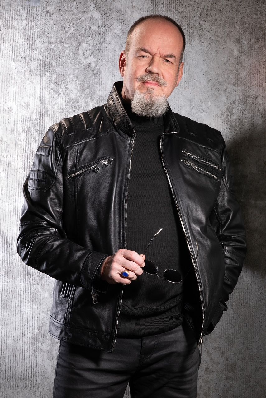 Eine Person mit Bart, die einen schwarzen Rollkragenpullover und eine Lederjacke trägt, posiert vor einem grauen, strukturierten Hintergrund.