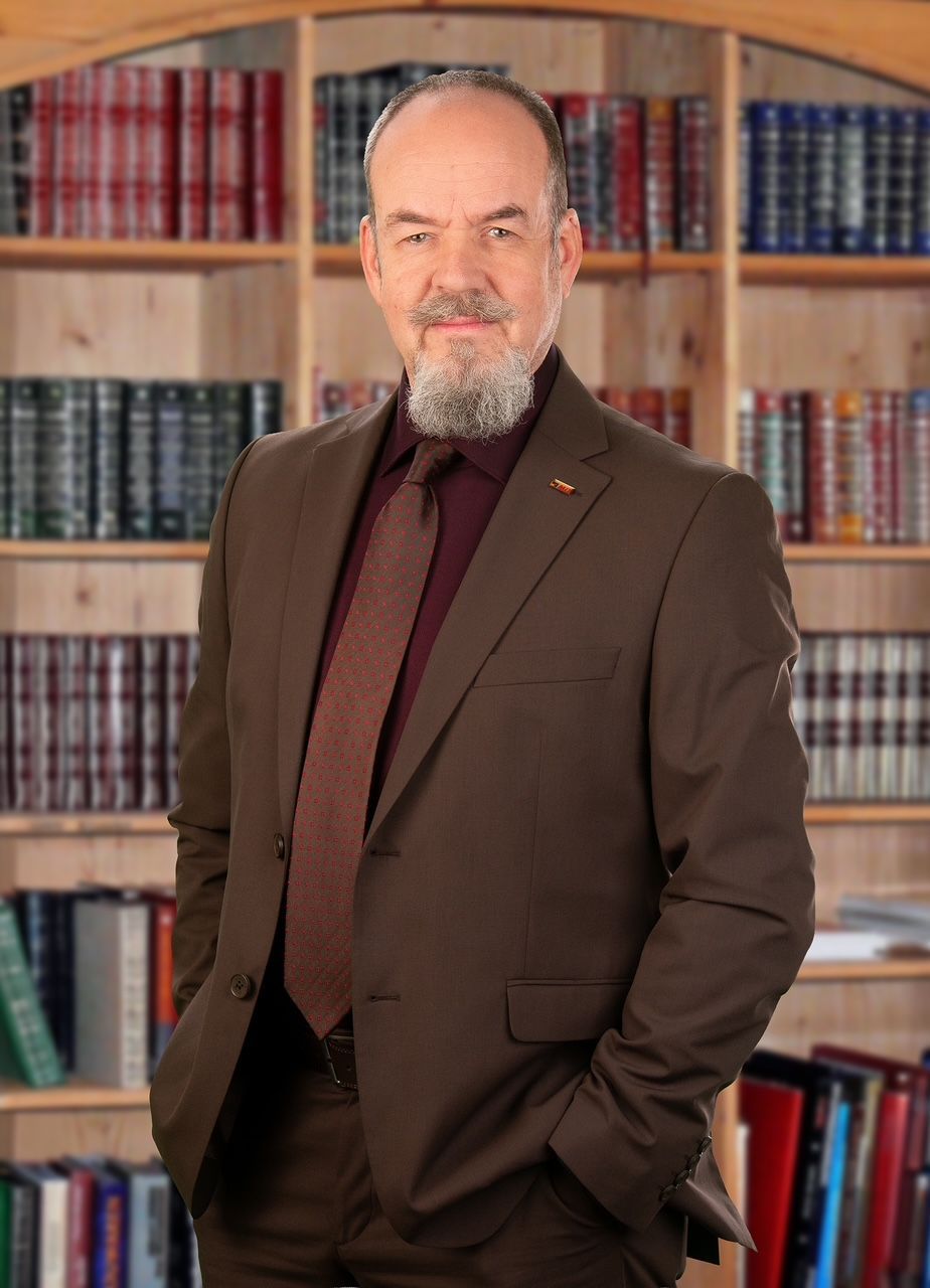 Ein professionelles Porträt eines Mannes in braunem Anzug und Krawatte, der mit den Händen in den Hosentaschen vor einem Bücherregal in einer Bibliothek steht.