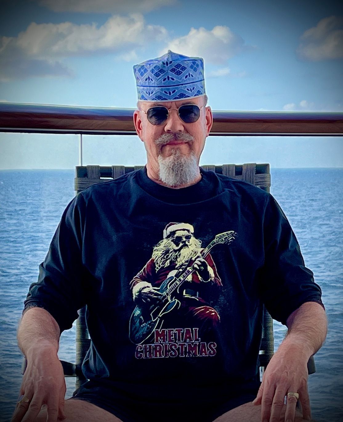 Eine Person mit blau gemustertem Hut, Sonnenbrille und schwarzem T-Shirt mit der Aufschrift „Metal Christmas“ sitzt vor einem Meerblick.