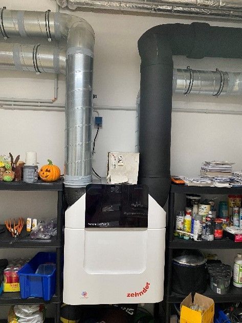 In einem Raum neben einem schwarzen Rohr steht eine weiße Maschine.
