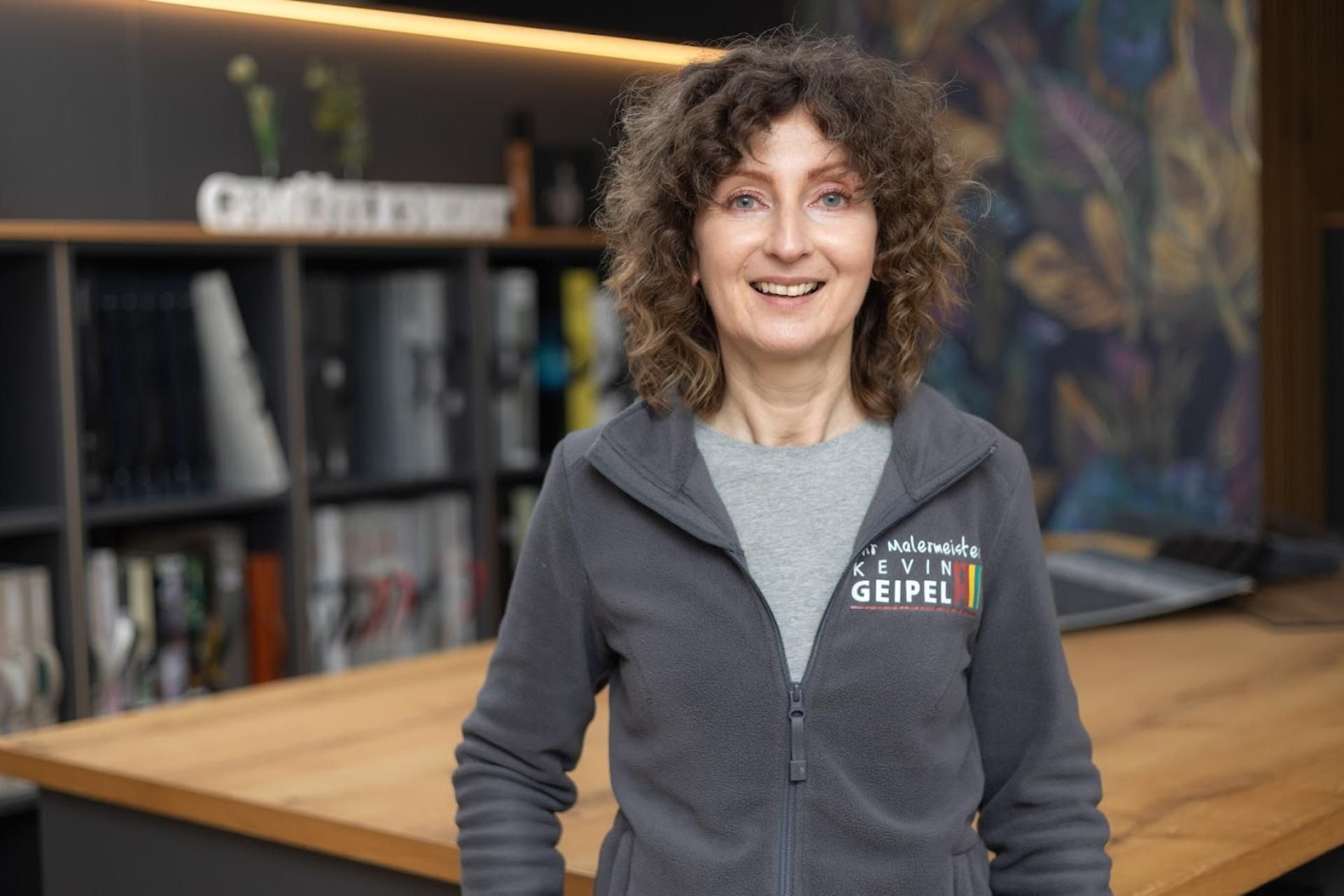 Eine Frau in einer grauen Jacke steht vor einem Holztisch.