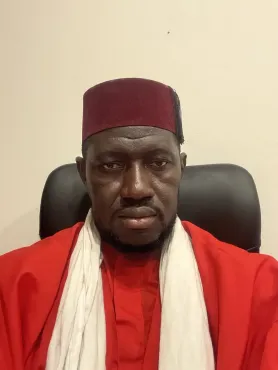 Hombre vestido con un traje rojo y una gorra granate, sentado en el interior, mirando directamente a la cámara.