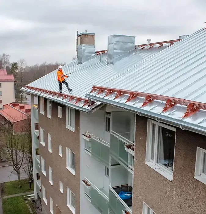 Oranssiin turvavarusteisiin pukeutunut työntekijä jyrkällä peltikatolla asentamassa kattoheloja kerrostaloon.