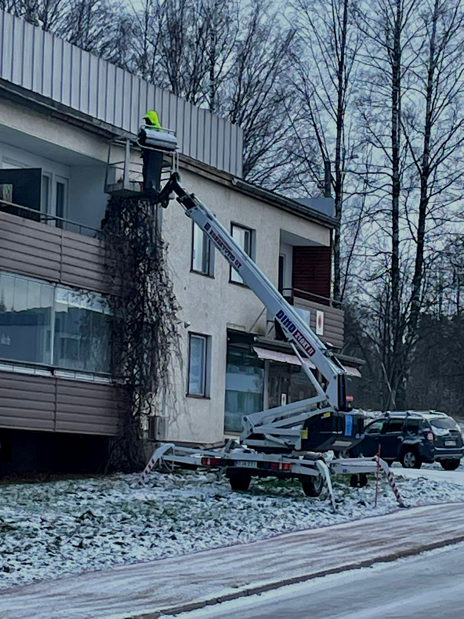 Työntekijä ämpärinosturilla leikkaamassa viiniköynnöksiä lumisen tien vieressä olevassa rakennuksessa