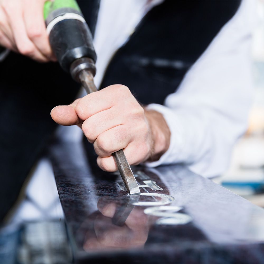 Henkilö käyttää sähköistä veistotyökalua kaivertaakseen kirjaimia kiillotettuun tummaan kivipintaan.