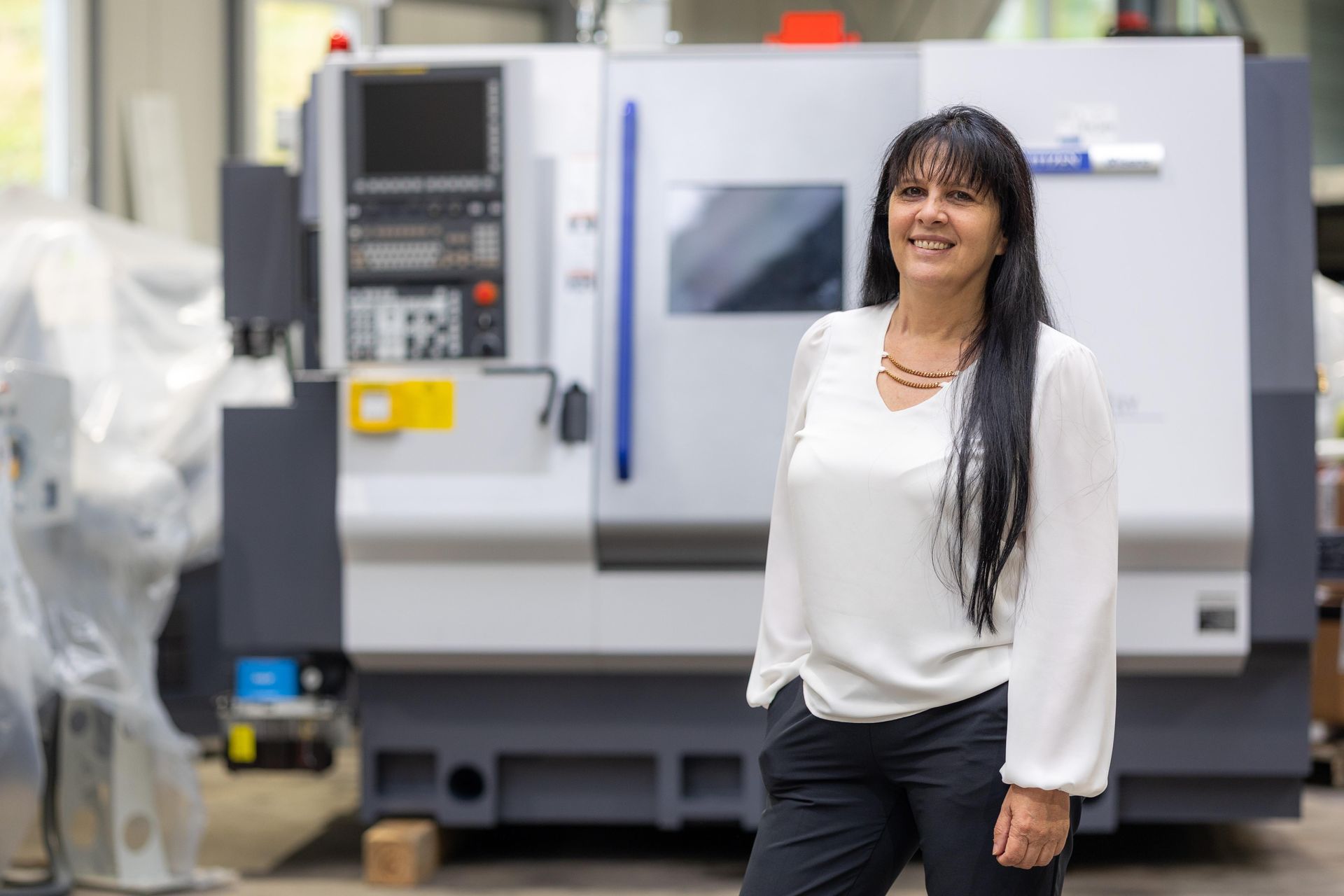 Eine lächelnde Frau steht neben einer CNC-Maschine in einer Werkstatt.