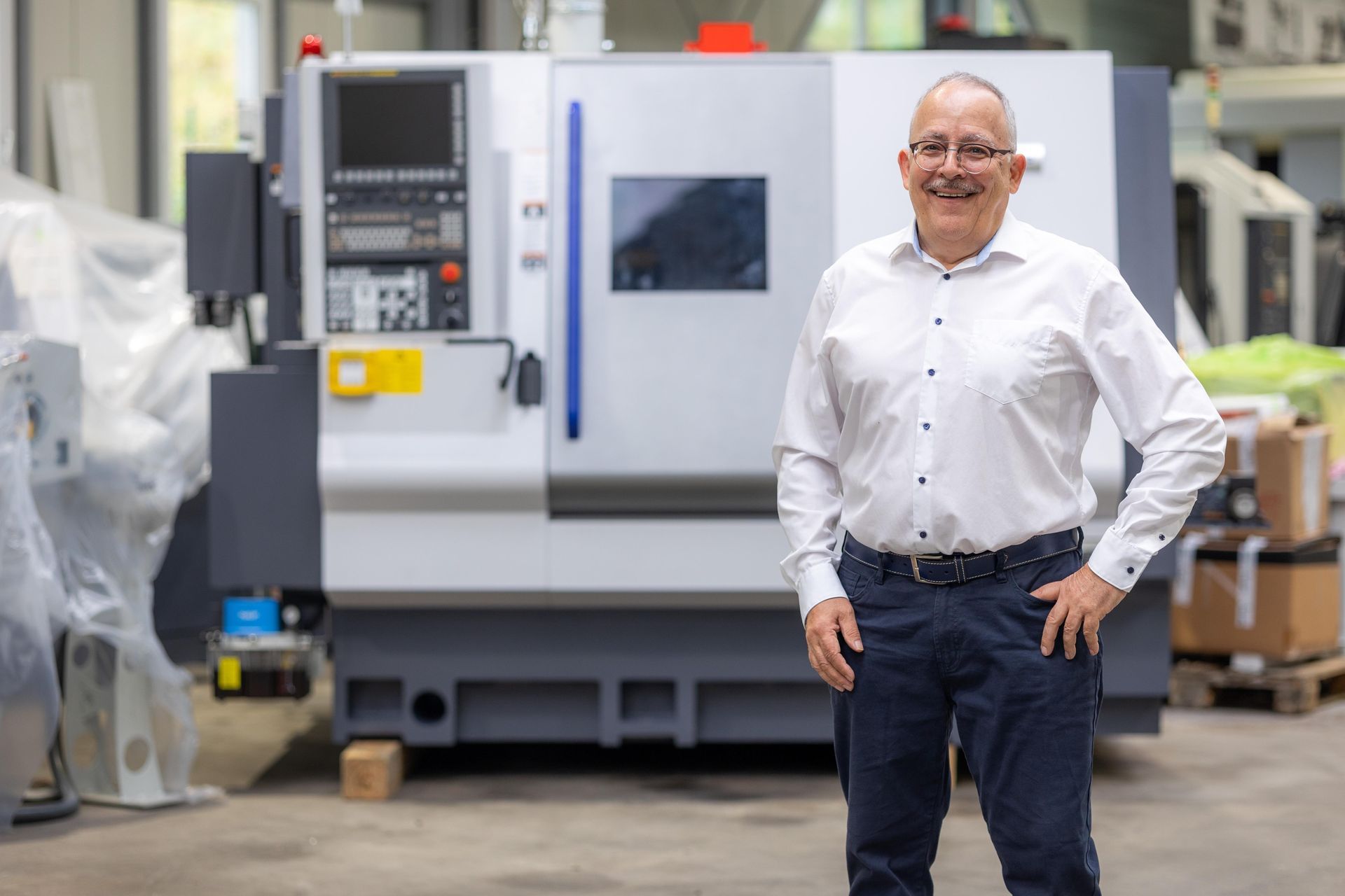 Ein Mann lächelt in einer Fabrik vor einer großen CNC-Maschine. Er trägt ein weißes Hemd und eine blaue Hose.