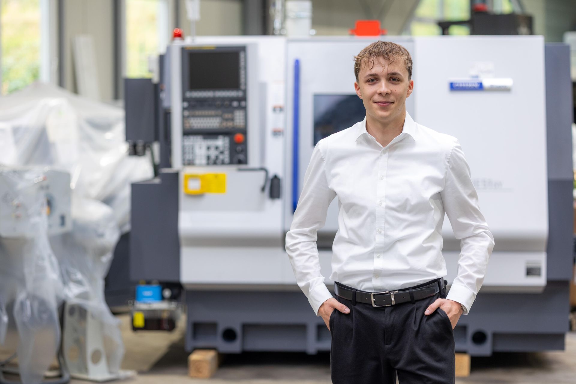 Mann in weißem Hemd und schwarzer Hose mit den Händen in den Taschen, steht vor einer CNC-Maschine.