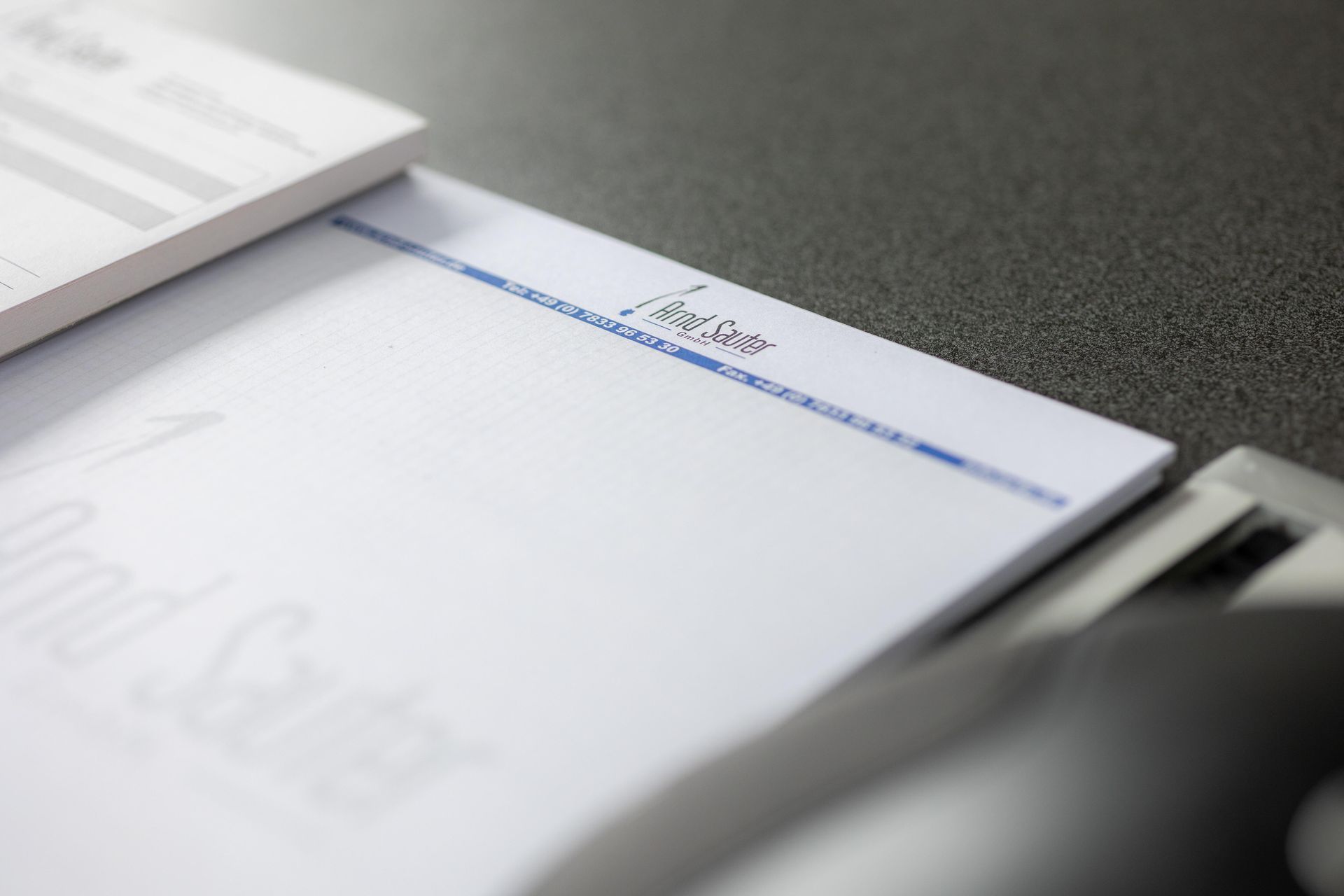 Briefpapier mit einem blauen Streifen und einem Logo auf einem Schreibtisch. Im Hintergrund ist ein Scheckbuch zu sehen.