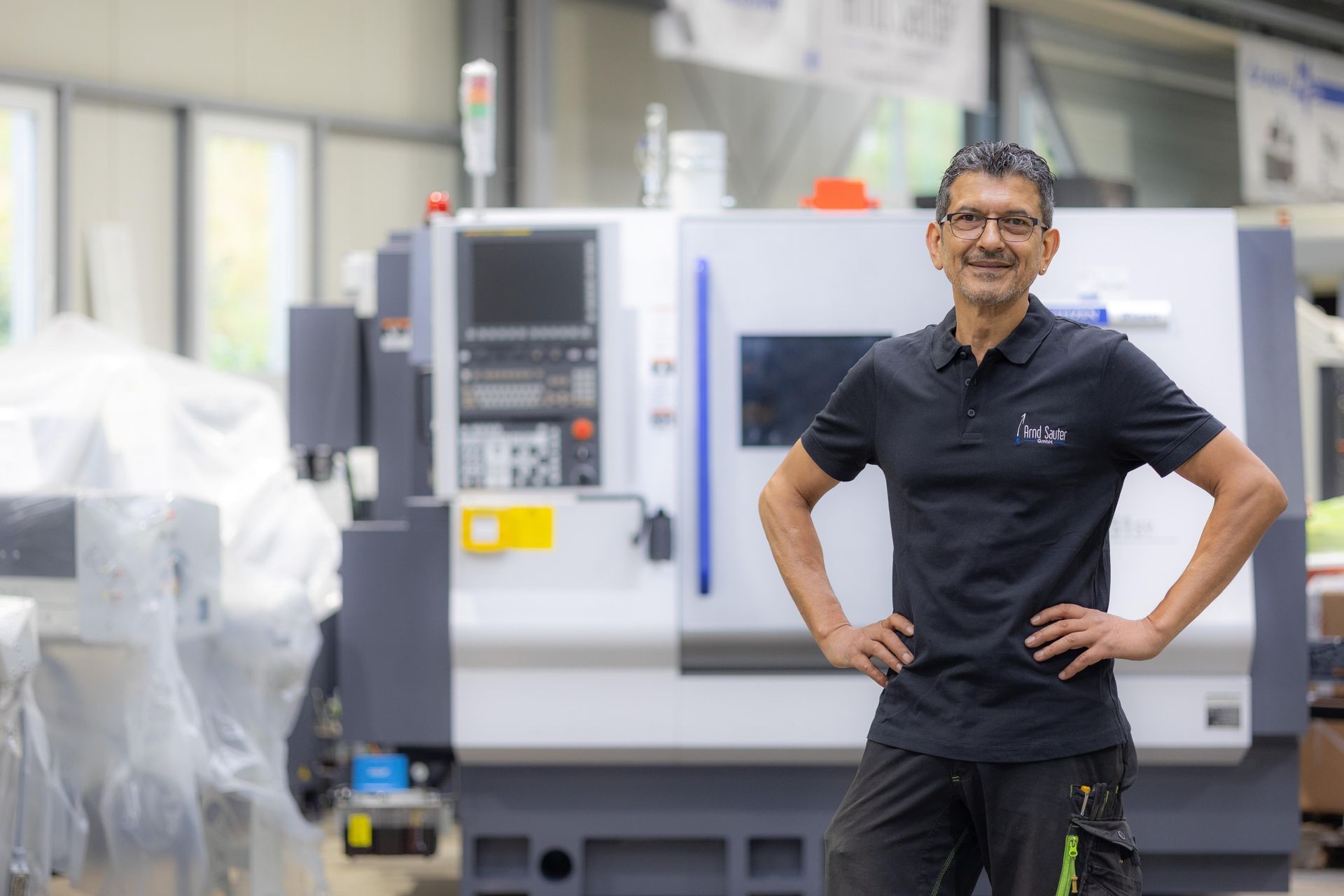 Ein Mann mit in die Hüften gestemmten Händen steht vor einer CNC-Maschine in einer Fabrikumgebung.