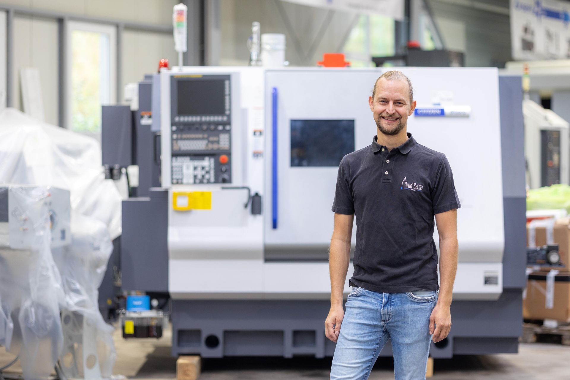 Ein Mann steht lächelnd vor einer Industriemaschine. Fabrikszene; er trägt ein schwarzes Hemd und Jeans.