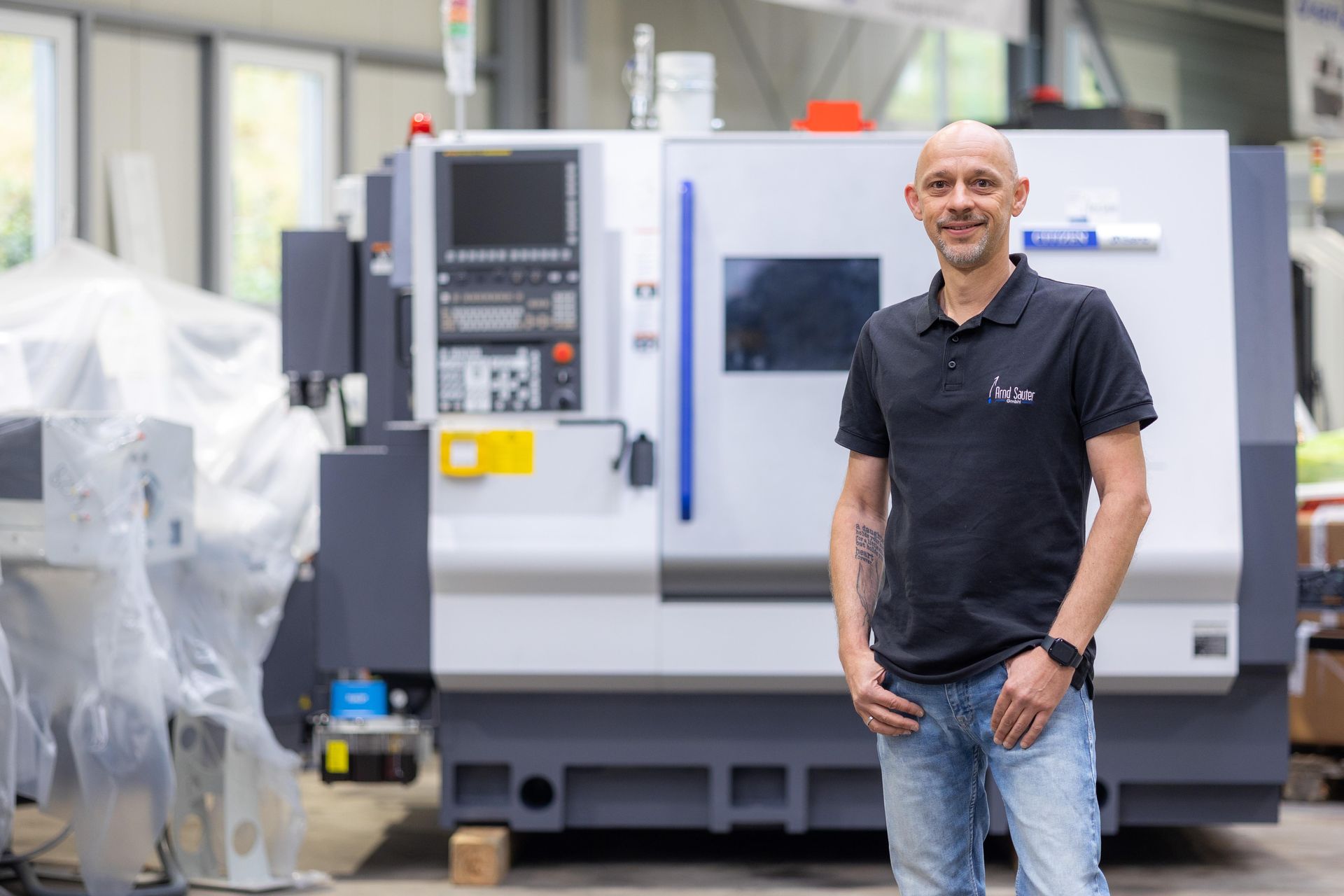 Ein Mann in einem schwarzen Hemd lächelt und posiert neben einer industriellen CNC-Maschine in einer Fabrik.