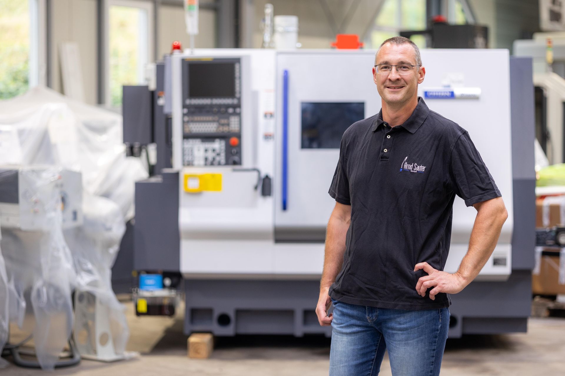 Ein Mann steht lächelnd vor einer CNC-Maschine. Er trägt ein schwarzes Hemd und Jeans und befindet sich in einer Werkstatt.