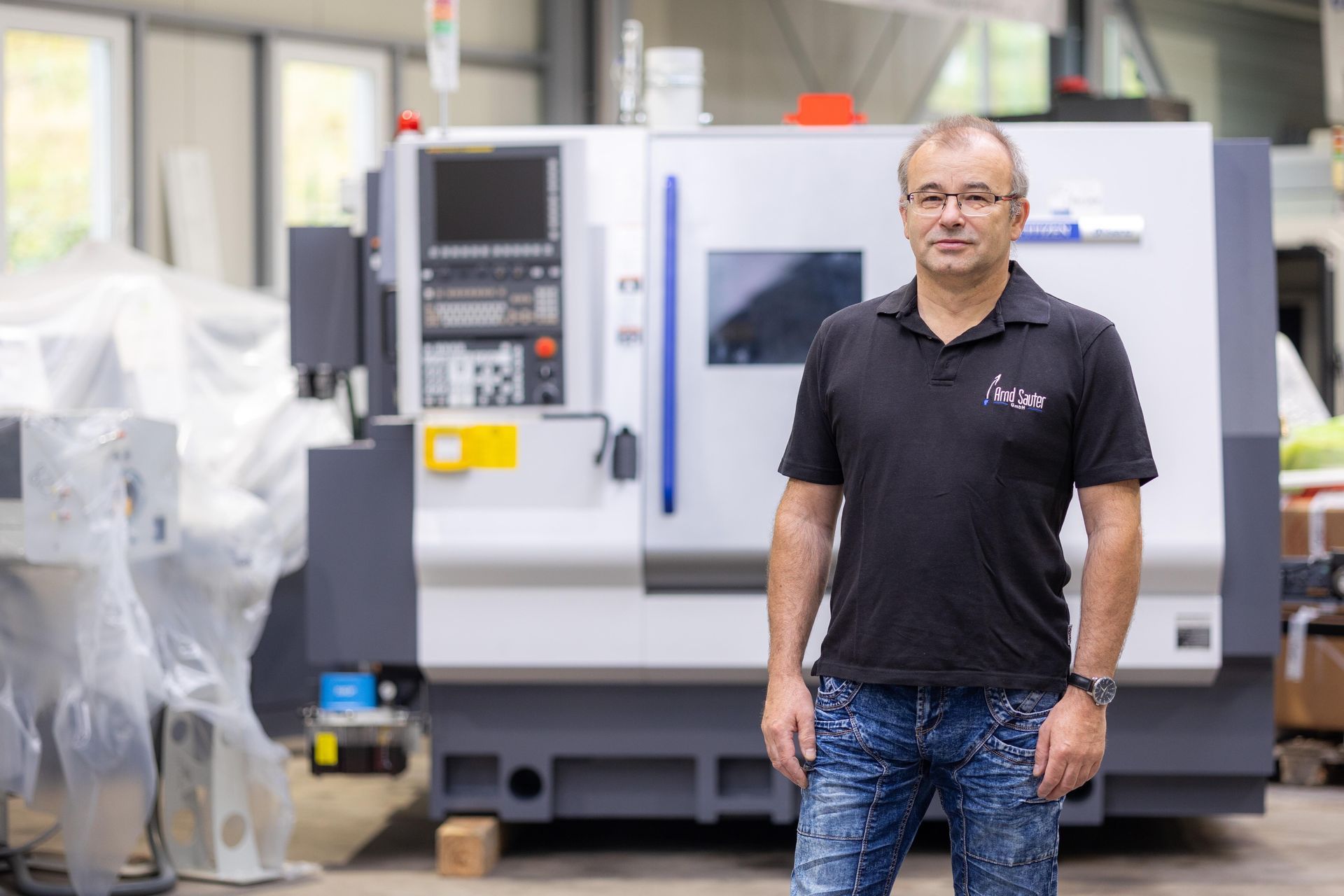 Ein Mann in schwarzem Hemd steht vor einer CNC-Maschine in einer Fabrikumgebung.