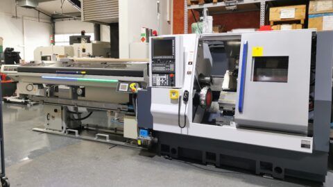 Eine große industrielle Drehmaschine in einer Fabrikhalle. Grau und blau, mit Bedienfeld und geöffneter Tür.