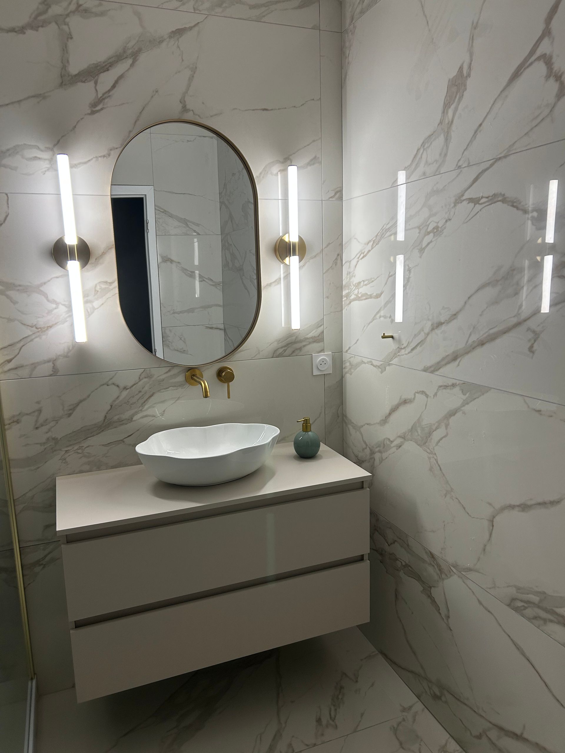 Meuble-lavabo moderne dans une salle de bains aux murs en marbre, avec miroir ovale, vasque à poser et appliques murales.