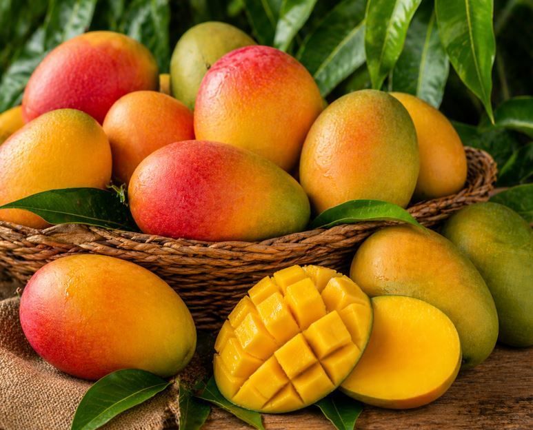 Cesta de mangos maduros con hojas verdes y rodajas de mango sobre una superficie de madera.