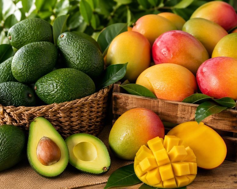 Aguacates, mangos y mango en rodajas sobre una mesa con cestas tejidas y hojas verdes.