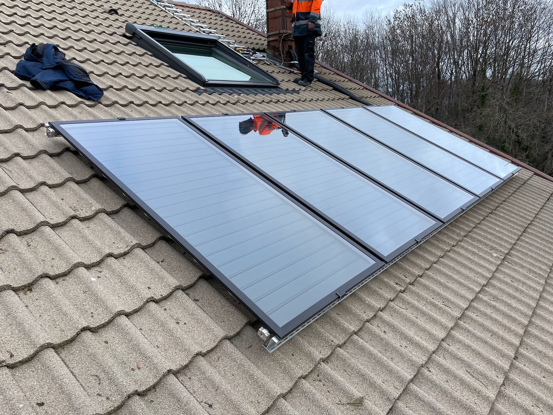 Installation de panneaux solaires bleus sur un toit de tuiles grises ; un ouvrier est visible près d'une fenêtre de toit