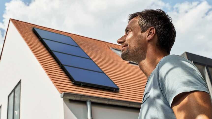 Un ouvrier en gilet orange et casque de chantier inspecte des panneaux solaires sur un toit sous un ciel bleu.