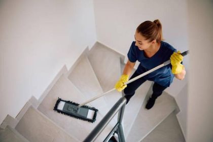 Eine Frau, die mit gelben Handschuhen eine graue Treppe wischt und dabei einen Wischmopp in der Hand hält, blickt nach unten.