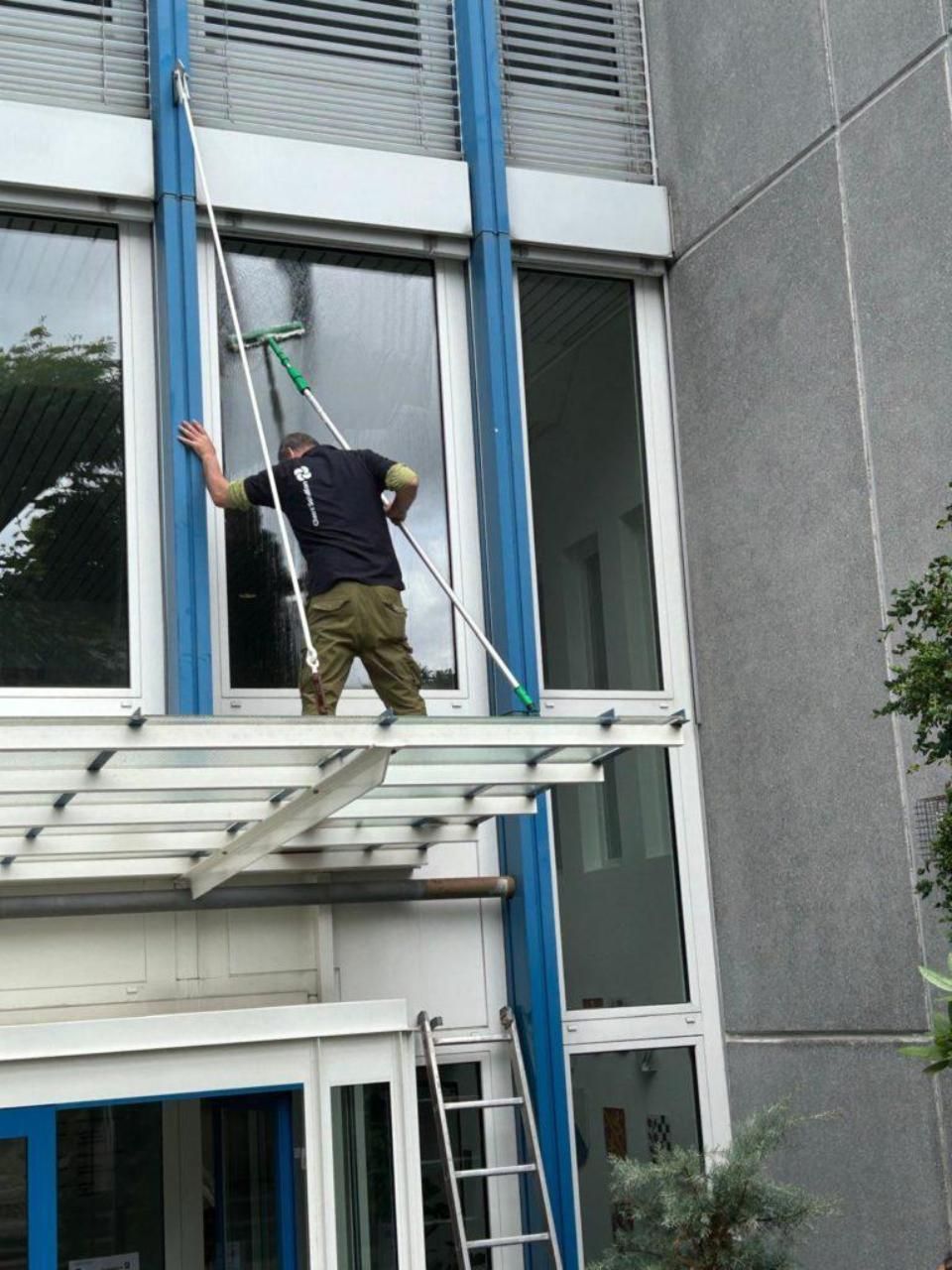 Ein Fensterputzer reinigt die Fenster eines Gebäudes. Er steht  auf einer Plattform und benutzt Abzieher mit langem Griff.