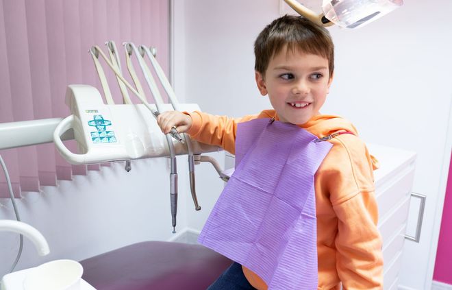 Un niño con una sudadera naranja y un babero dental morado sonríe en un sillón de dentista con instrumentos dentales cerca.