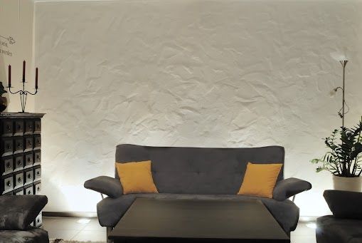 Graues Sofa mit gelben Kissen vor strukturierter weißer Wand, flankiert von Möbeln und Pflanzen.