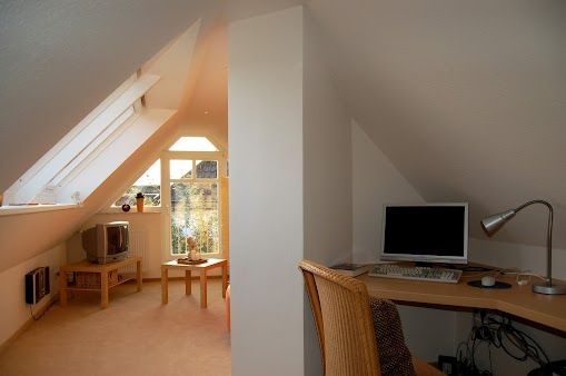 Dachzimmer mit Dachschräge, Schreibtisch mit Computer, Stuhl und kleiner Sitzecke mit Fenster und Fernseher.