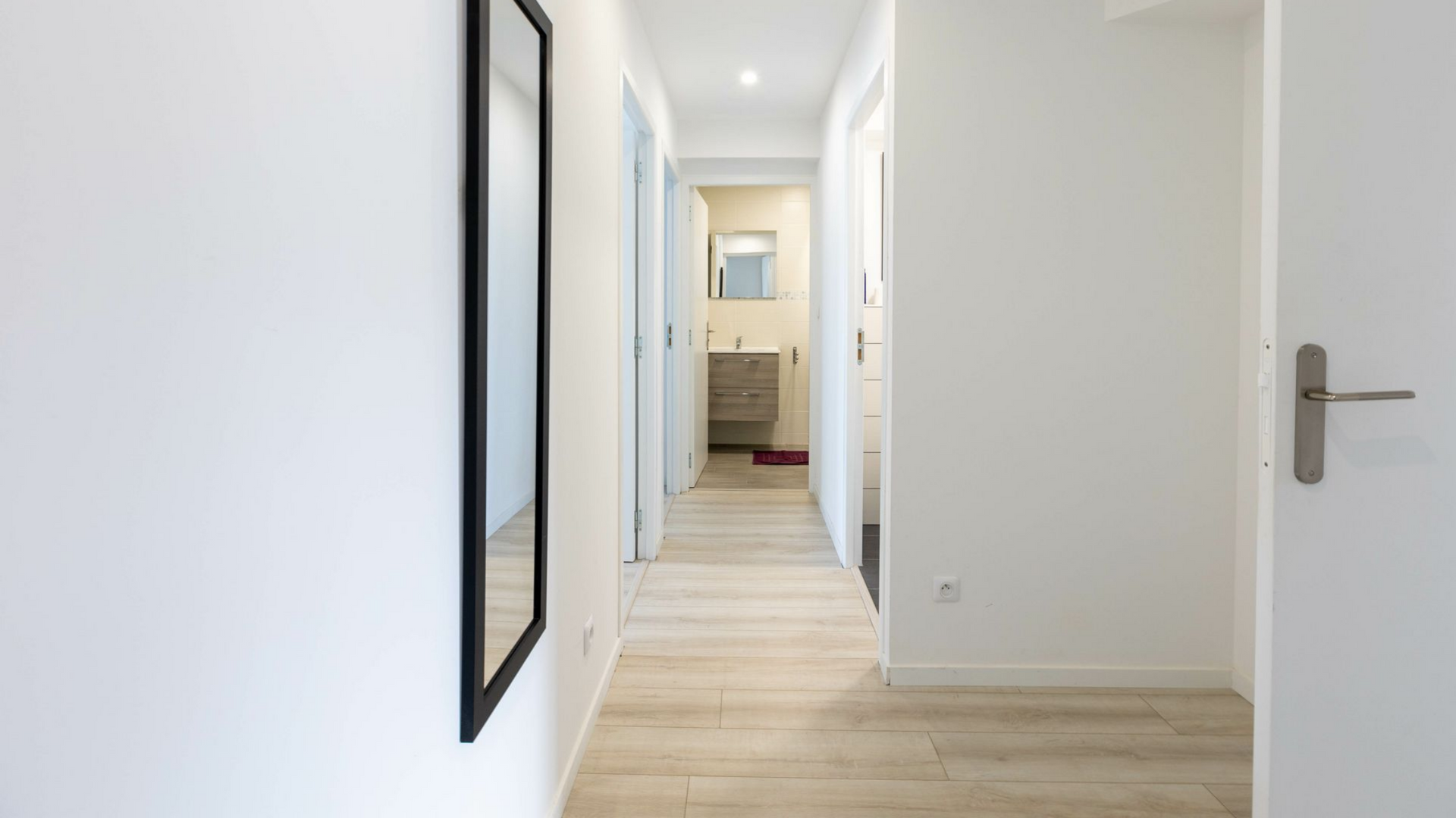Couloir lumineux avec murs blancs, parquet clair et vue sur une salle de bains au fond.