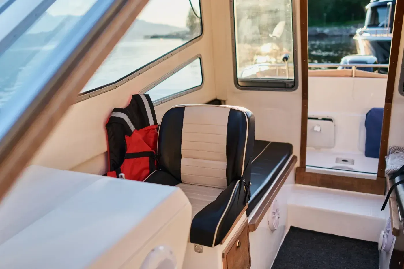 Vista interior de la cabina de un barco con silla de capitán, chaleco salvavidas y ventanas con vista al agua.