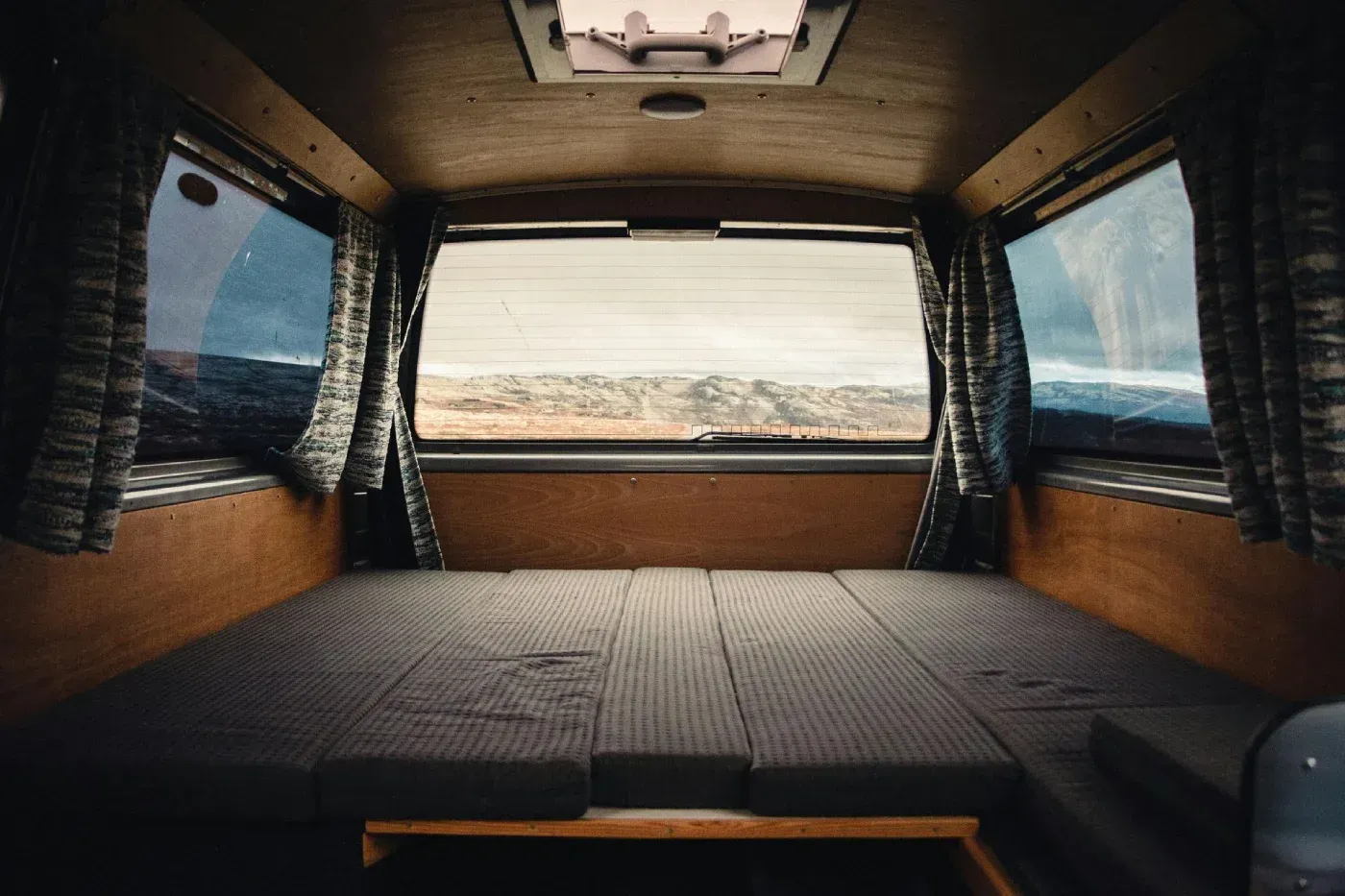 Interior de una furgoneta con cama y ventanas con vistas a un paisaje; paneles marrones, cortinas azules.